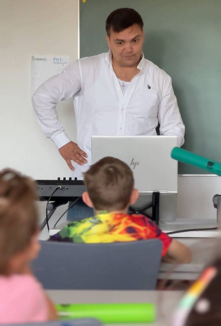 Ein Lehrer steht hinter einem Klavier und einem Laptop vor den Kindern im Klassenraum während einer Stunde der frühen Musikerziehung.