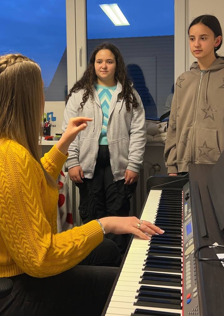 Eine Lehrerin sitzt am Klavier und erklärt etwas zwei Schülerinnen, die daneben stehen. Gesangsunterricht.