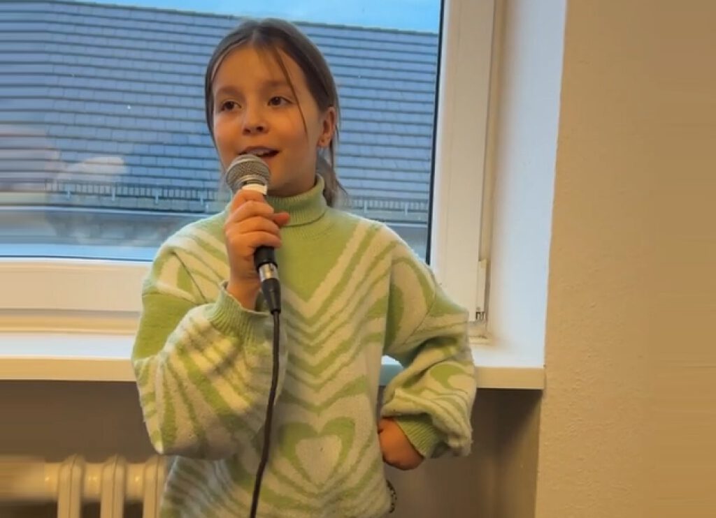 Ein Mädchen singt in ein Mikrofon und steht vor einem Fenster.