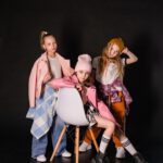 Drei Mädchen posieren vor schwarzem Hintergrund, eine sitzt auf einem Stuhl, zwei stehen daneben.