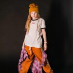 Ein Mädchen in weißem T-Shirt und orangefarbener Hose posiert vor schwarzem Hintergrund.