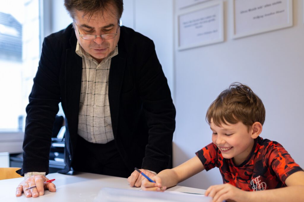 Ein Lehrer betreut einen Jungen beim Schreiben von Notenübungen, der Junge lacht fröhlich.