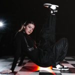 Танцовщица в чёрной одежде сидит на полу и поднимает ногу вверх.