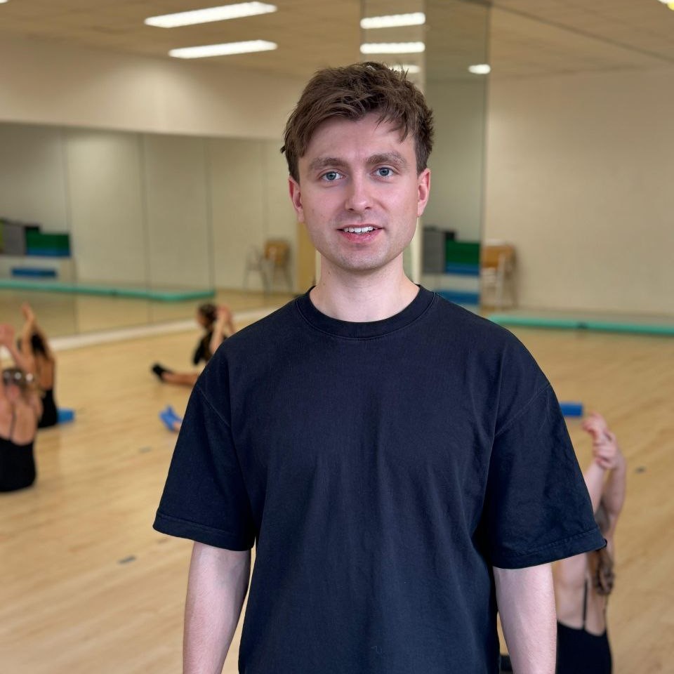 Ein Mann in einem schwarzen T-Shirt lächelt. Hinter ihm machen Kinder Gymnastik in einem Saal mit Spiegeln.