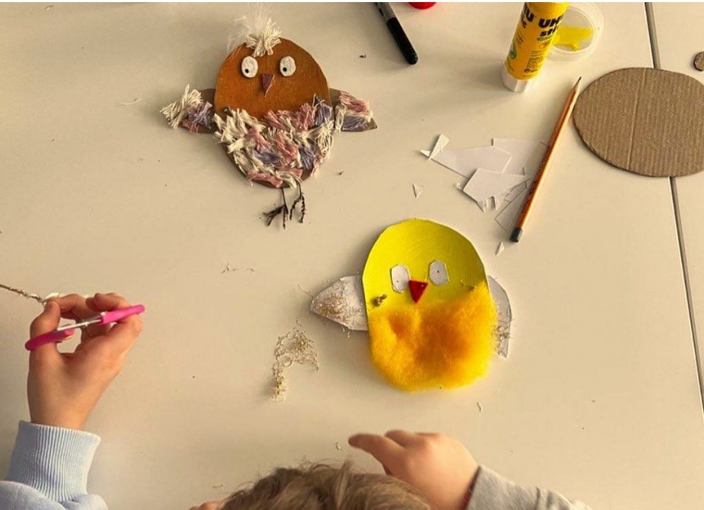 Ein Kind schneidet mit einer Schere Material für eine Bastelarbeit aus. Auf dem Tisch liegen Applikationen in Form von Küken, Kleber, ein Bleistift und Pappstücke