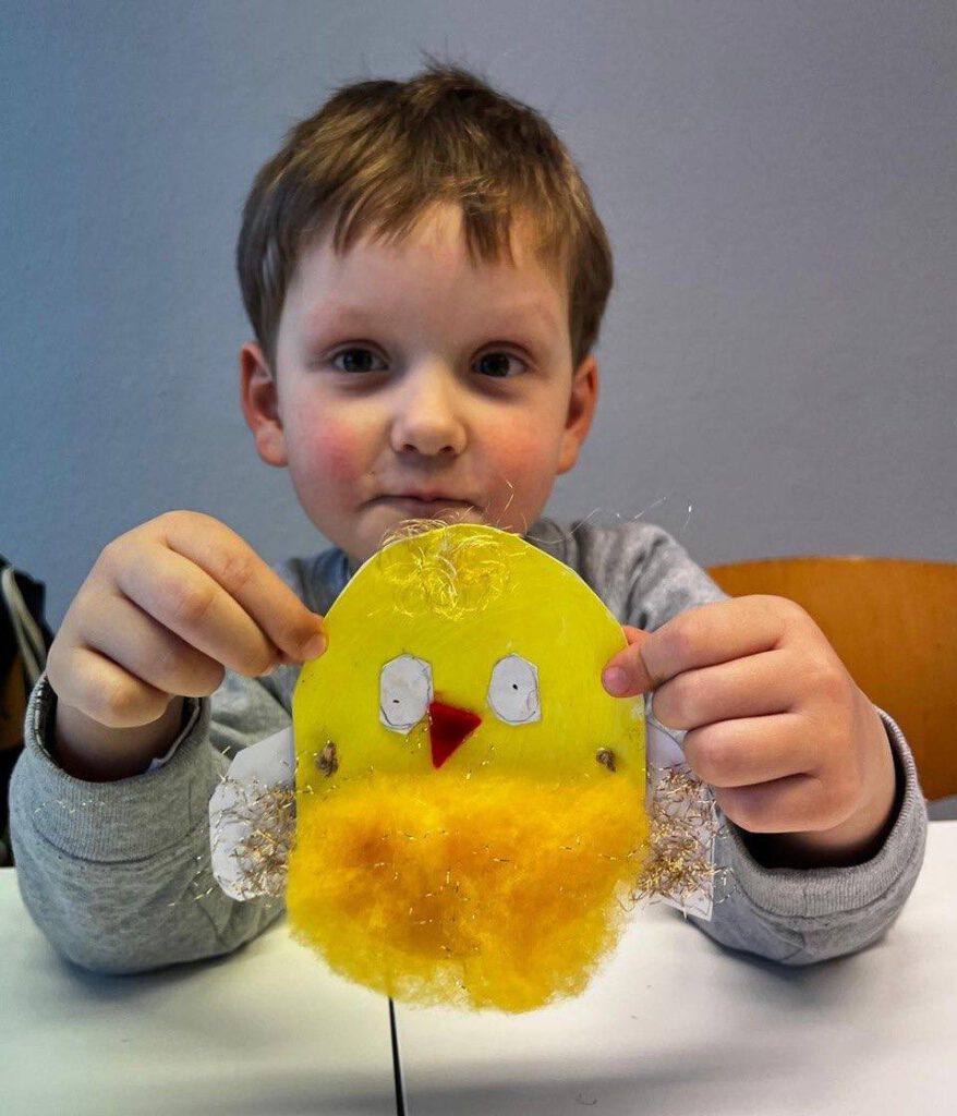 Мальчик сидит за столом и держит в руках сделанную им аппликацию жёлтого птенца с красным клювом.