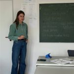 Преподаватель объясняет грамматику у доски; на стене висят плакаты.