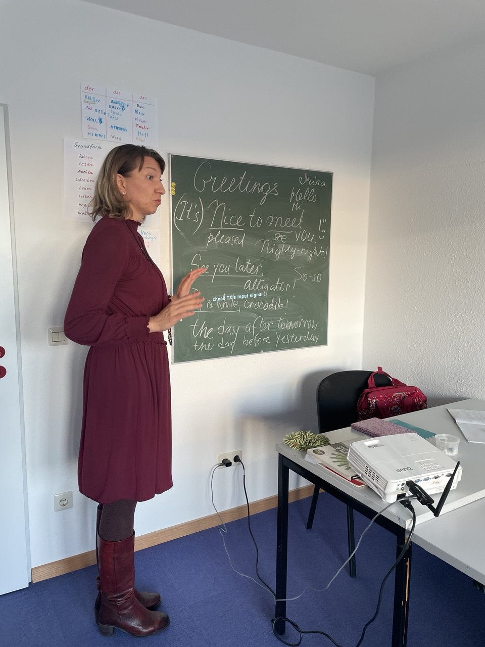 Englischlehrerin steht an der Tafel „Greetings“ und erklärt Redewendungen.