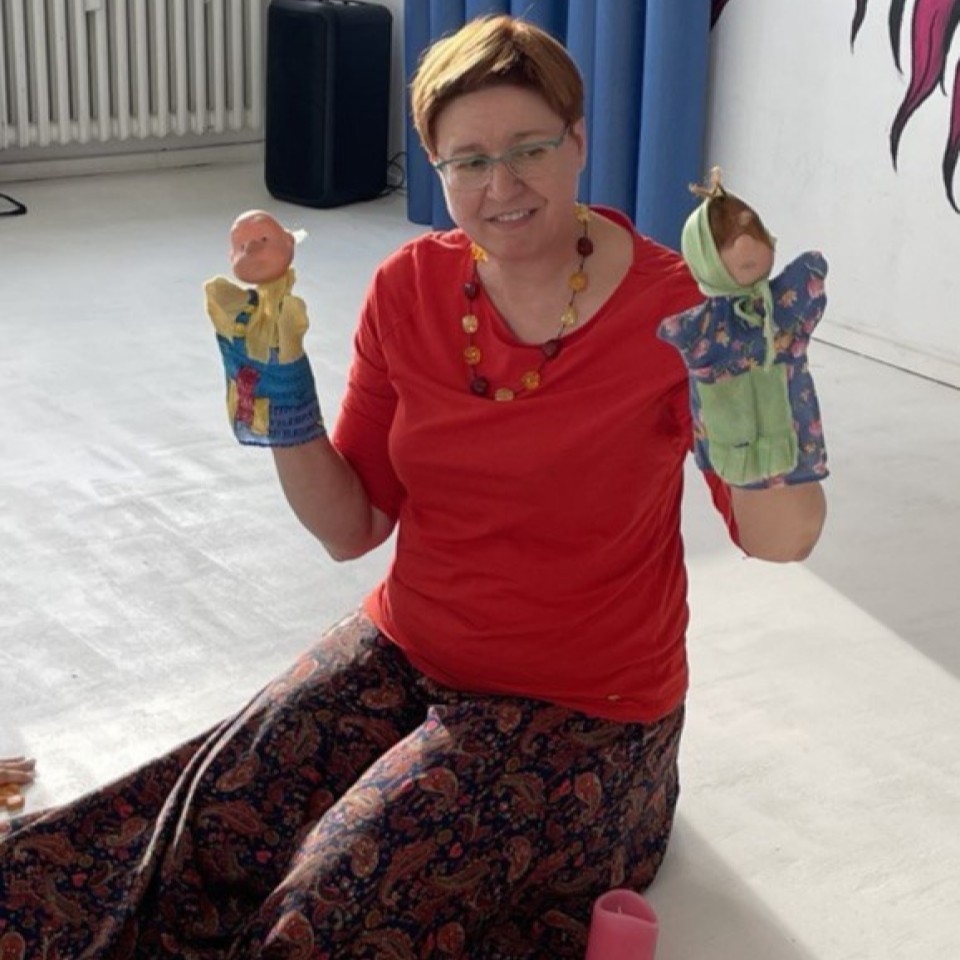 Eine Frau lächelt und sitzt auf dem Boden. Sie trägt Theaterpuppen an ihren Händen.