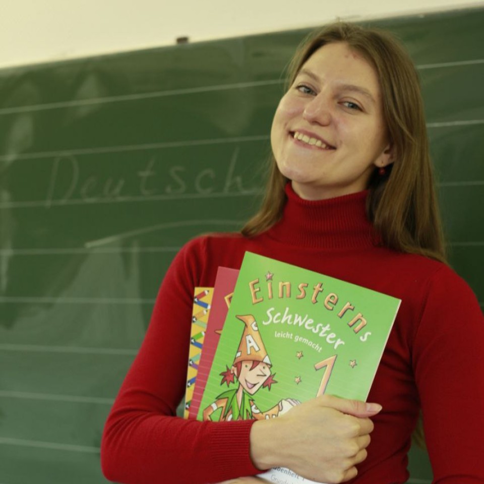 Eine Frau lächelt neben einer Tafel. In ihren Händen hält sie Lehrbücher.