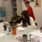 Zwei Mädchen zeichnen am Tisch, die Lehrerin im roten Pullover beobachtet und hilft. An der Wand hängen Gemälde.