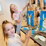 Zwei Mädchen malen an Staffeleien Bilder mit blauem Hintergrund, eine lächelt fröhlich.