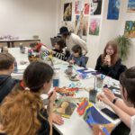 Ältere Jugendliche zeichnen am großen Tisch, die Lehrerin mit Hut hilft und beobachtet. An den Wänden hängen bunte Bilder.