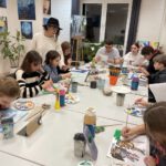 Jugendliche sitzen an den Tischen und malen, die Lehrerin steht daneben und berät. Im Atelier hängen viele farbenfrohe Bilder.