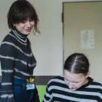 Eine Schülerin zeichnet mit einem schwarzen Marker auf Papier, die Lehrerin steht daneben und lächelt.