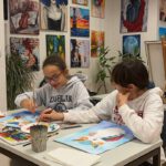 Zwei Mädchen sitzen am Tisch und malen mit Aquarellfarben. An der Wand hängen viele farbenfrohe Bilder.