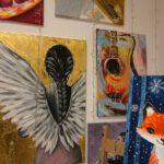 Mehrere farbenfrohe Gemälde hängen an der Wand: ein Engel mit Flügeln, eine Gitarre, ein Fuchs und abstrakte Kompositionen.
