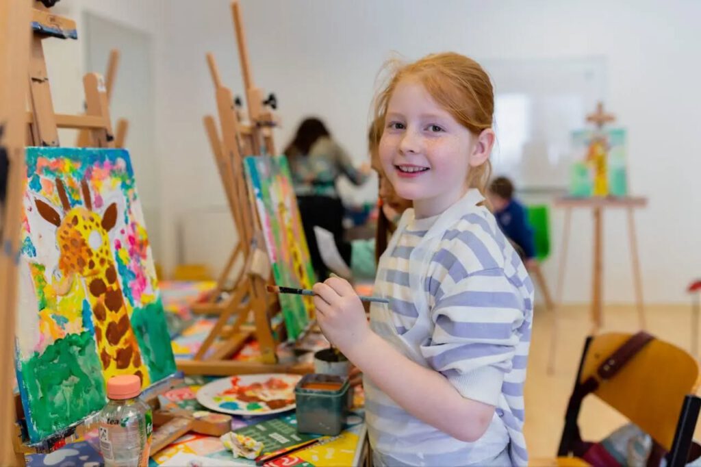 Ein Mädchen lächelt mit einem Pinsel in der Hand, neben ihr steht ihr farbenfrohes Bild mit einer Giraffe auf der Staffelei.