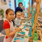 Mehrere Mädchen und die Lehrerin malen an Staffeleien und lächeln in die Kamera.