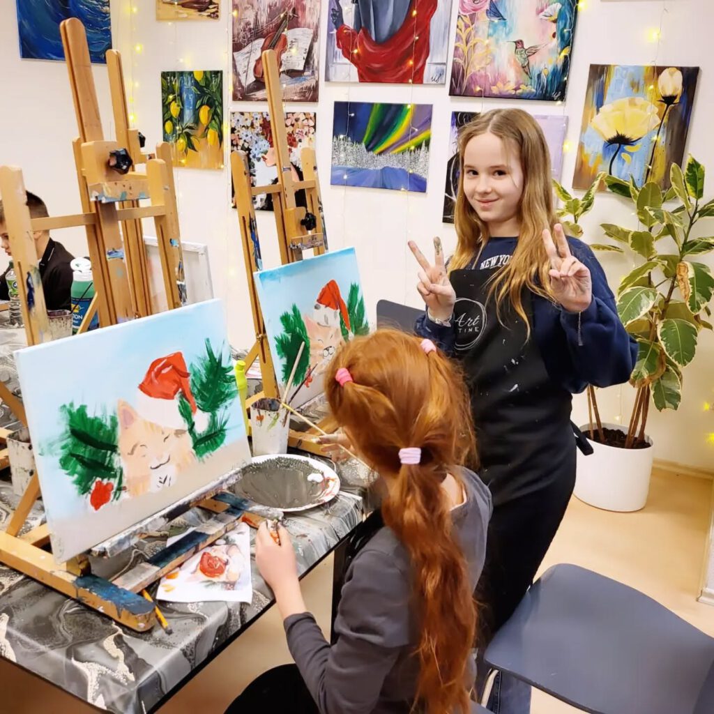 Zwei Mädchen malen eine Katze mit roter Mütze, eine zeigt das Victory-Zeichen und lächelt.