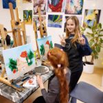 Zwei Mädchen malen eine Katze mit roter Mütze, eine zeigt das Victory-Zeichen und lächelt.