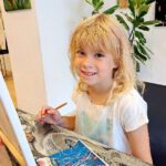 Ein blondes Mädchen sitzt an der Staffelei mit einem Pinsel in der Hand und lächelt, daneben liegt ihr Bild.