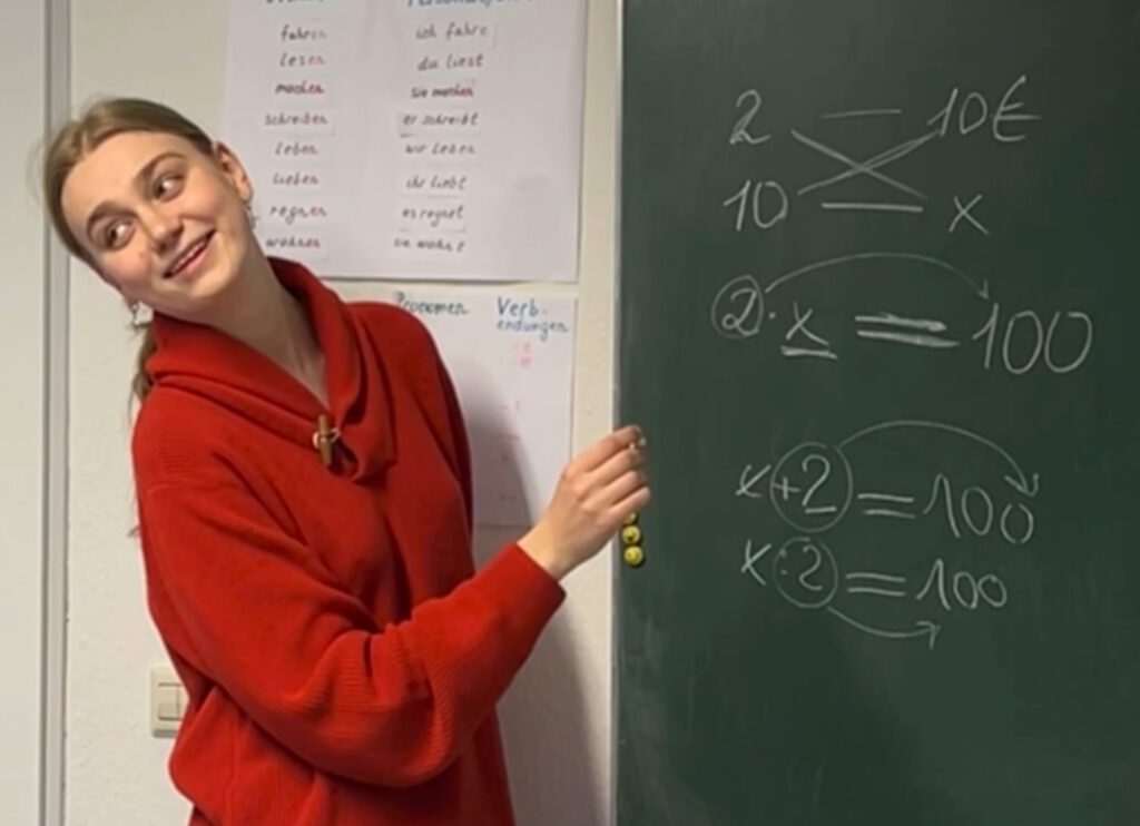 Lehrerin im roten Pullover erklärt am Tafeleintrag Proportions-/Prozentrechnen.