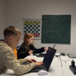 Jugendliche programmieren an Laptops; an der Tafel steht ‚Hello World‘ mit Pfeildiagramm.