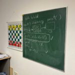Tafel im Kurs: ‚Hello World‘ und einfache Programmlogik; daneben Schach-Lernposter.