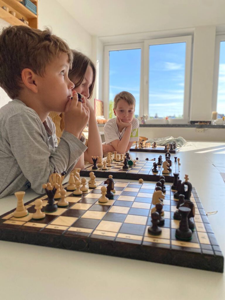 Drei Kinder analysieren Positionen an zwei Schachbrettern in einem hellen Raum mit großen Fenstern.