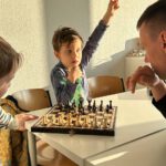 Kind hebt die Hand, um einen Zug zu besprechen; gegenüber sitzt der Trainer am Schachbrett.