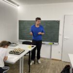 Lehrer erläutert an der Tafel und am Brett eine Stellung; Schülerin folgt der Erklärung.