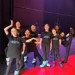 Юные танцовщицы в футболках с логотипом позируют за кулисами.