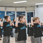 Девочки в зале выполняют синхронное танцевальное упражнение с поднятыми руками.