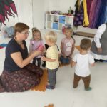 Педагог протягивает малышу стаканчик; несколько детей стоят кругом на ковриках.