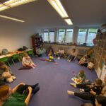 Круговое занятие для малышей и родителей: взрослые и дети сидят на полу большим кругом и играют с развивающими игрушками в светлой комнате.