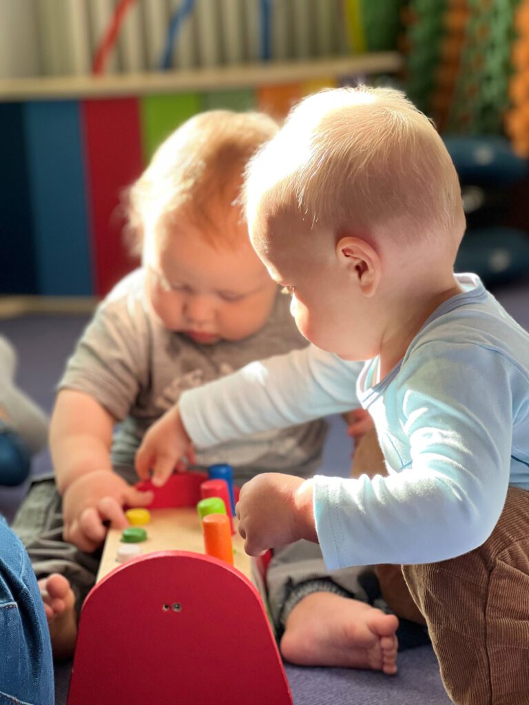 Zwei Babys spielen im Sonnenlicht mit einem Holz-Steckspiel; Fokus auf Feinmotorik.