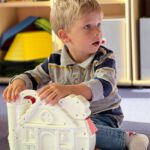 Малыш сидит на полу и рассматривает большую игрушку-дом; на заднем плане — стеллажи с книгами.