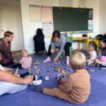 Родители и дети сидят кругом; малыши играют с погремушками, мячиками и палочками, педагог задаёт ритм.