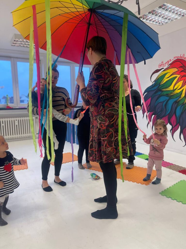 Bewegungsspiel mit großem Regenbogenschirm und Bändern; Kleinkinder und Eltern stehen im Kreis.