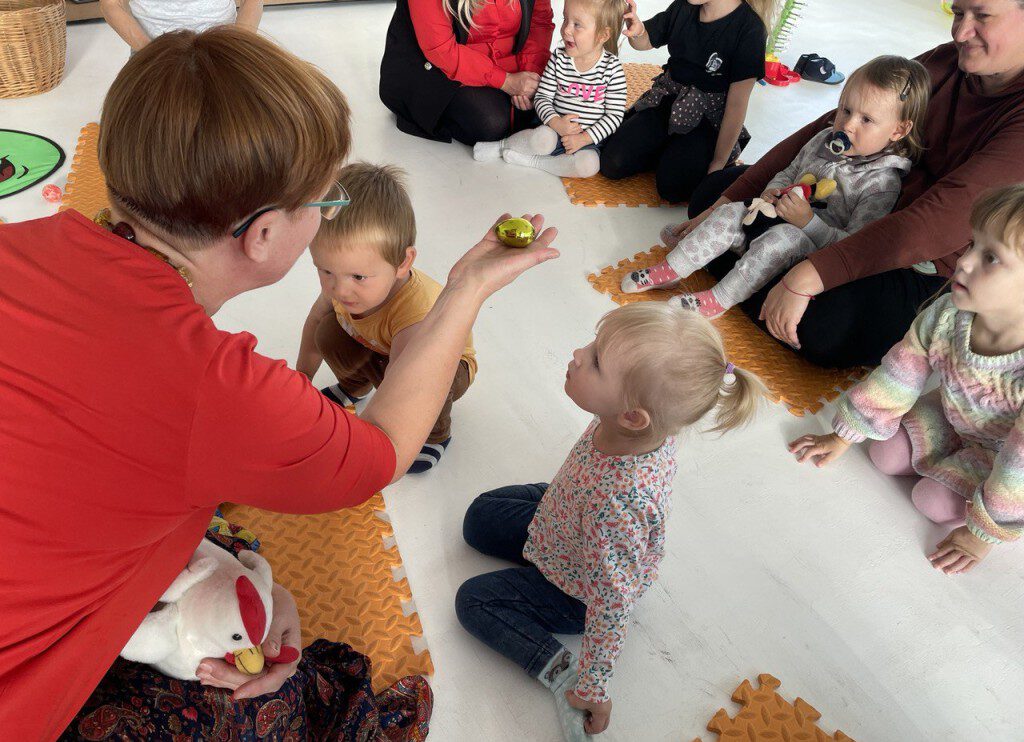 Pädagogin zeigt einem Kleinkind eine kleine Figur; Kinder sitzen im Halbkreis auf Matten.