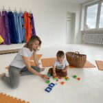 Мама и малыш сортируют цветные кубики на полу; рядом плетёная корзина.