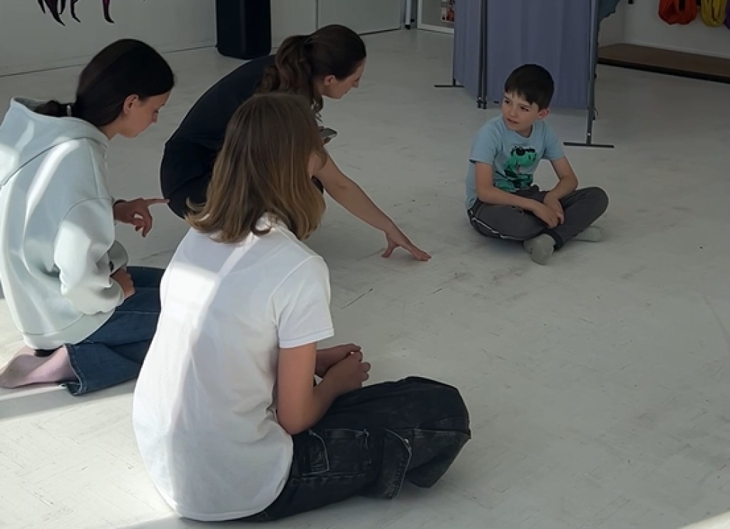 Theatercoach spricht mit einem Jungen, zwei Jugendliche hören am Boden zu – Impro-Übung zu Interaktion und Zuhören.