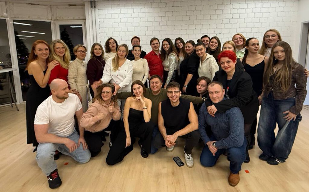 Gruppenfoto von Teilnehmerinnen und Teilnehmern in einem hellen Raum mit großen Fenstern. Alle stehen und sitzen fröhlich zusammen, lächeln in die Kamera.