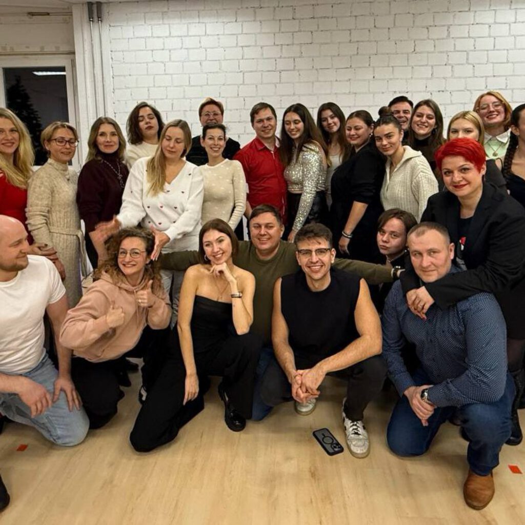 Групповое фото участников в светлом зале с большими окнами. Все стоят и сидят вместе, улыбаются в камеру.